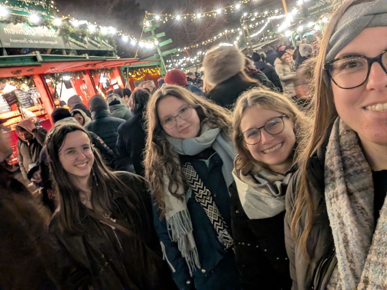 Vier Frauen mit Brillen und Winterkleidung stehen lächelnd zusammen, mit einer Menschenmenge und festlichen Lichtern im Hintergrund.