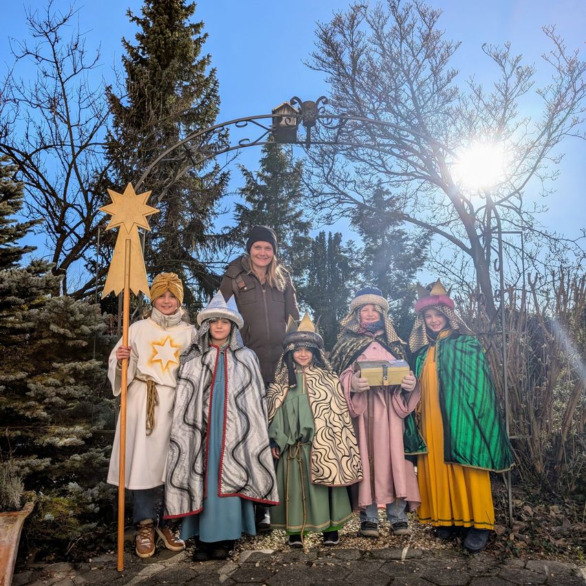 Eine Gruppe von Kindern in mittelalterlichen Kostümen, die Requisiten halten und unter einem Sternenbogen stehen, mit einer lächelnden Frau im Hintergrund.