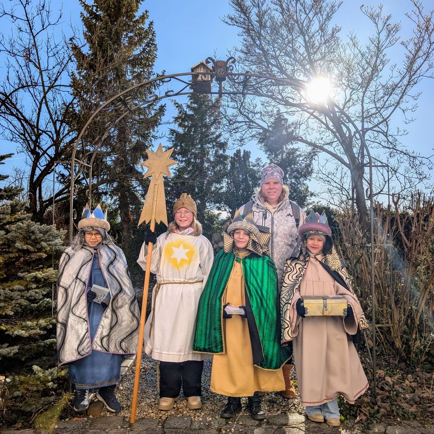 Fünf Personen in verschiedenen Kostümen posieren für ein Foto in einem Garten, wobei eine Frau einen Stern hält und ein Mann mit Hut in der Mitte steht.