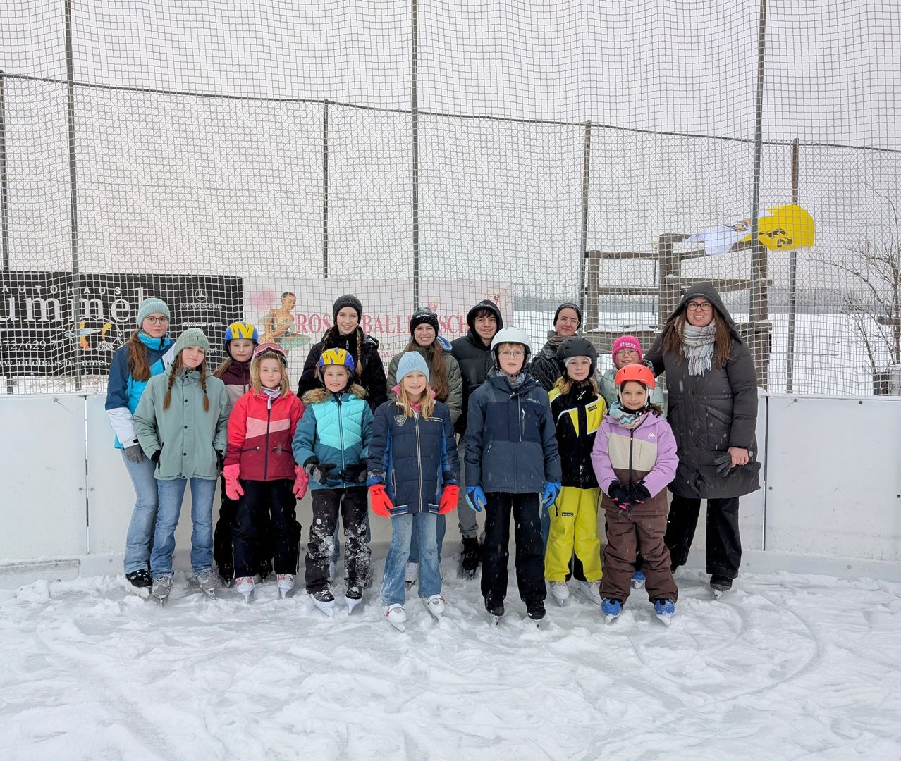 Eine Gruppe von Kindern und Erwachsenen in Winterkleidung und Helmen posiert für ein Foto im Schnee vor einem Zaun und einem Gebäude.