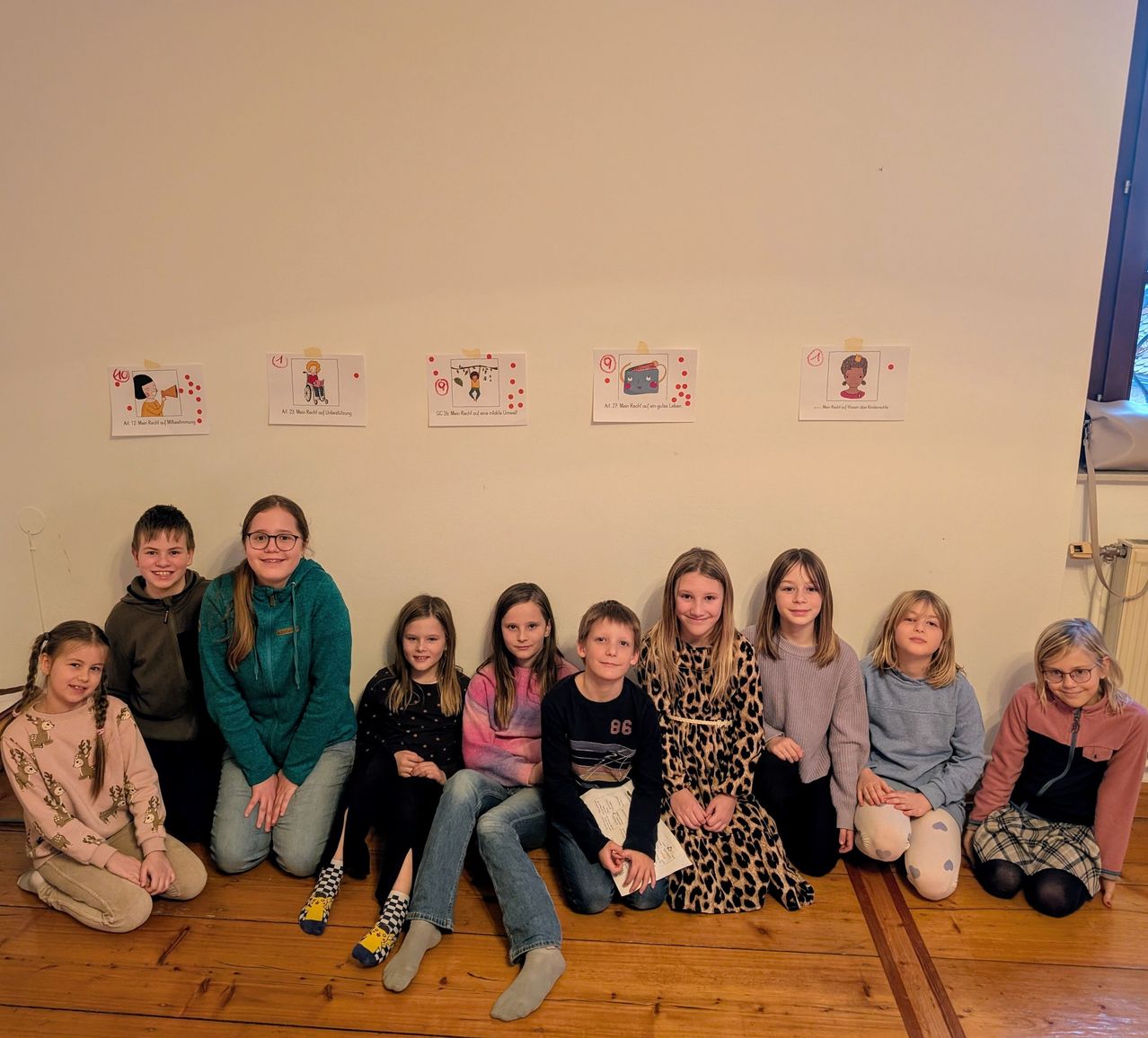 A group of children sit on the floor in front of a wall with drawings. They are all smiling and appear to be posing for a photograph. The drawings on the wall seem to depict different activities.