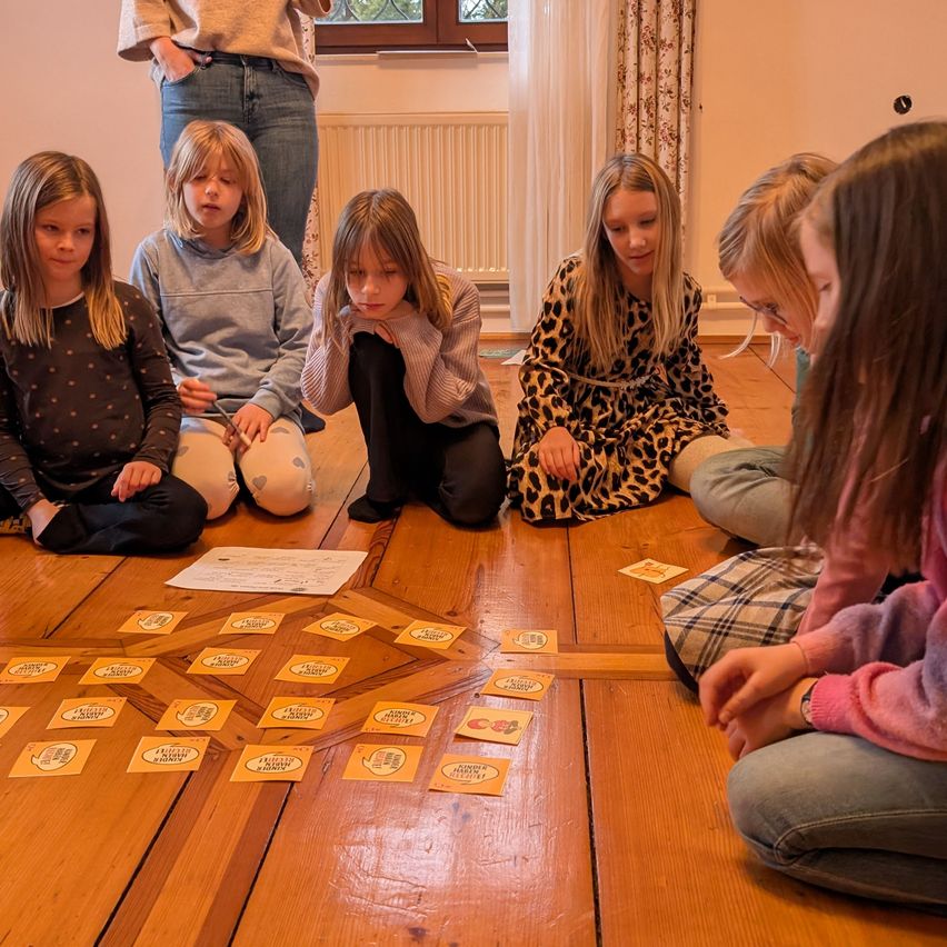 A group of girls sit in a circle on the floor playing a board game with cards. One girl looks at the floor. Another person stands behind them.