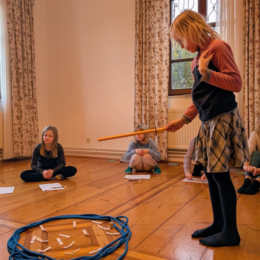 A girl stands and holds a long stick with multiple small cards attached to it, while other children sit on the floor and hold papers.