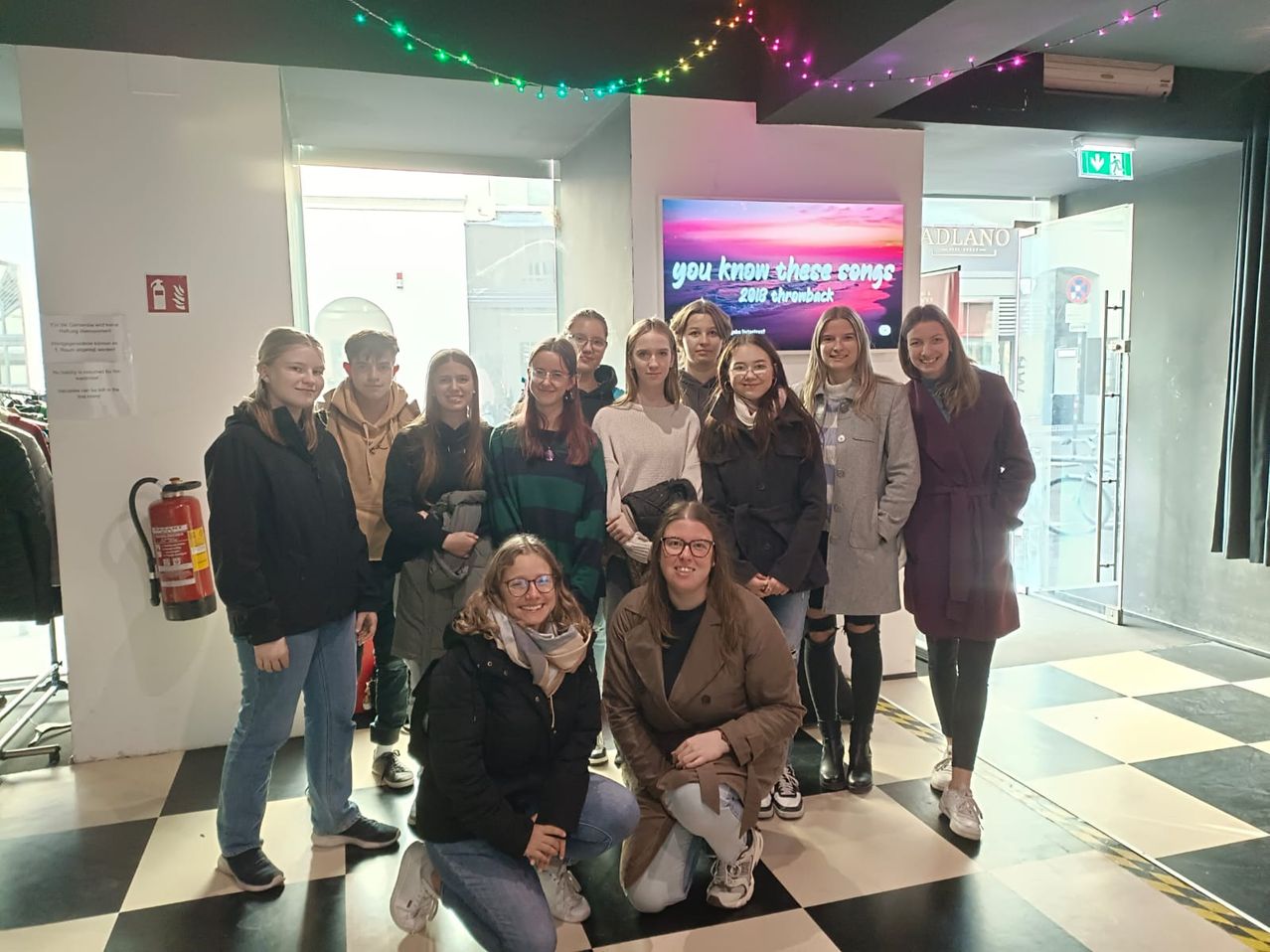 Eine Gruppe junger Frauen posiert für ein Foto in einer Lobby, mit einem Monitor hinter ihnen, der eine Nachricht anzeigt. Sie lächeln alle und tragen Winterkleidung.
