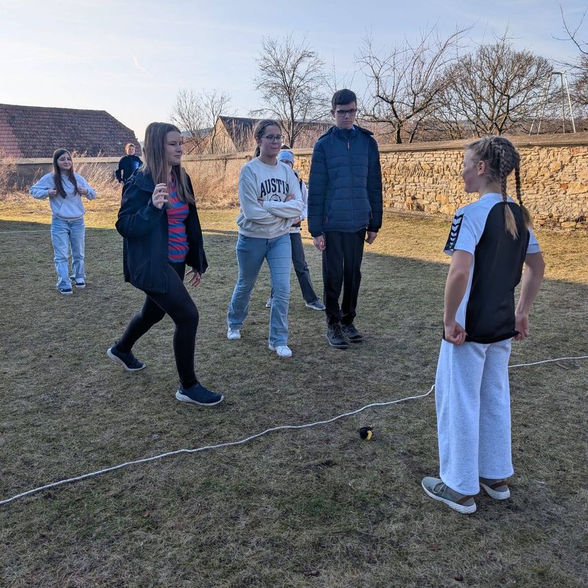 A group of people are standing on a grassy field, possibly in a park, wearing casual clothing. They are probably doing some exercise.