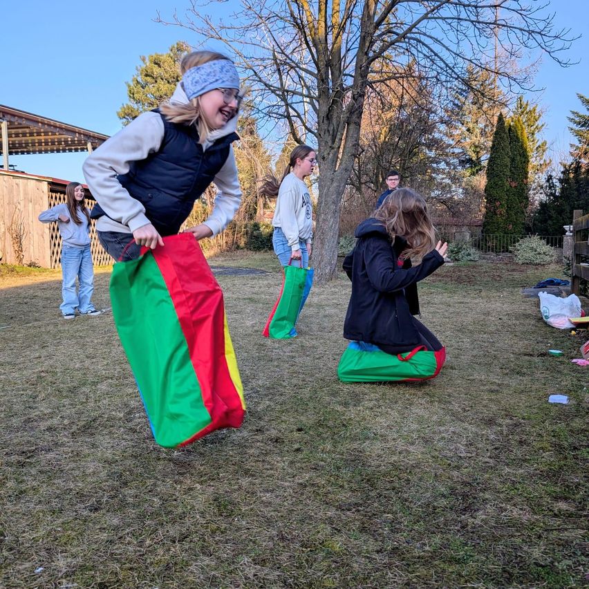 Several kids are playing a game of sack race outside. One girl is jumping in the air with a colorful sack. Another girl is kneeling on the ground with her sack. The other kids are watching. Behind them are trees and a fence.