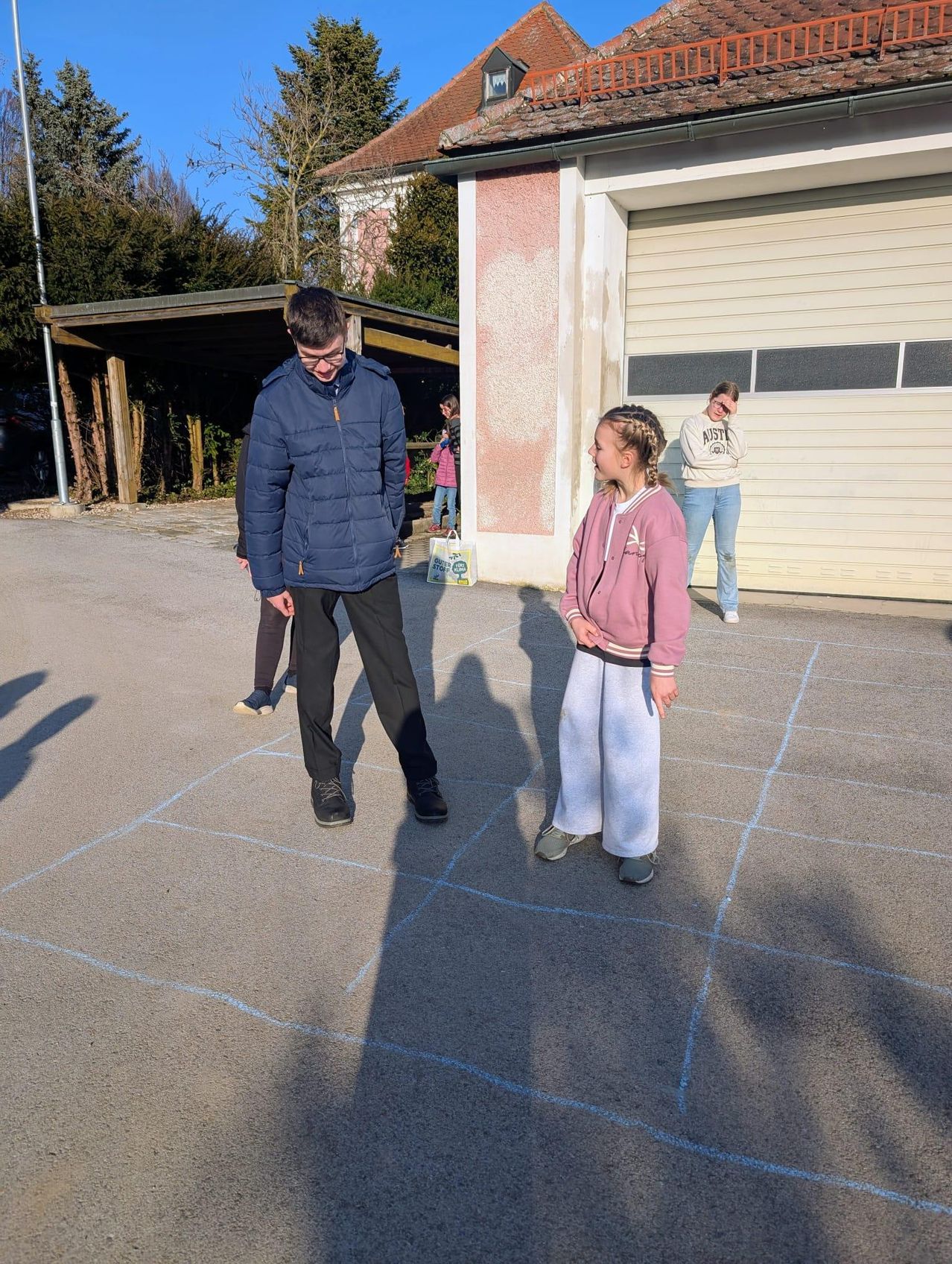 A man and a girl play hopscotch on a concrete court, with a woman standing behind them. Behind them is a garage with trees.