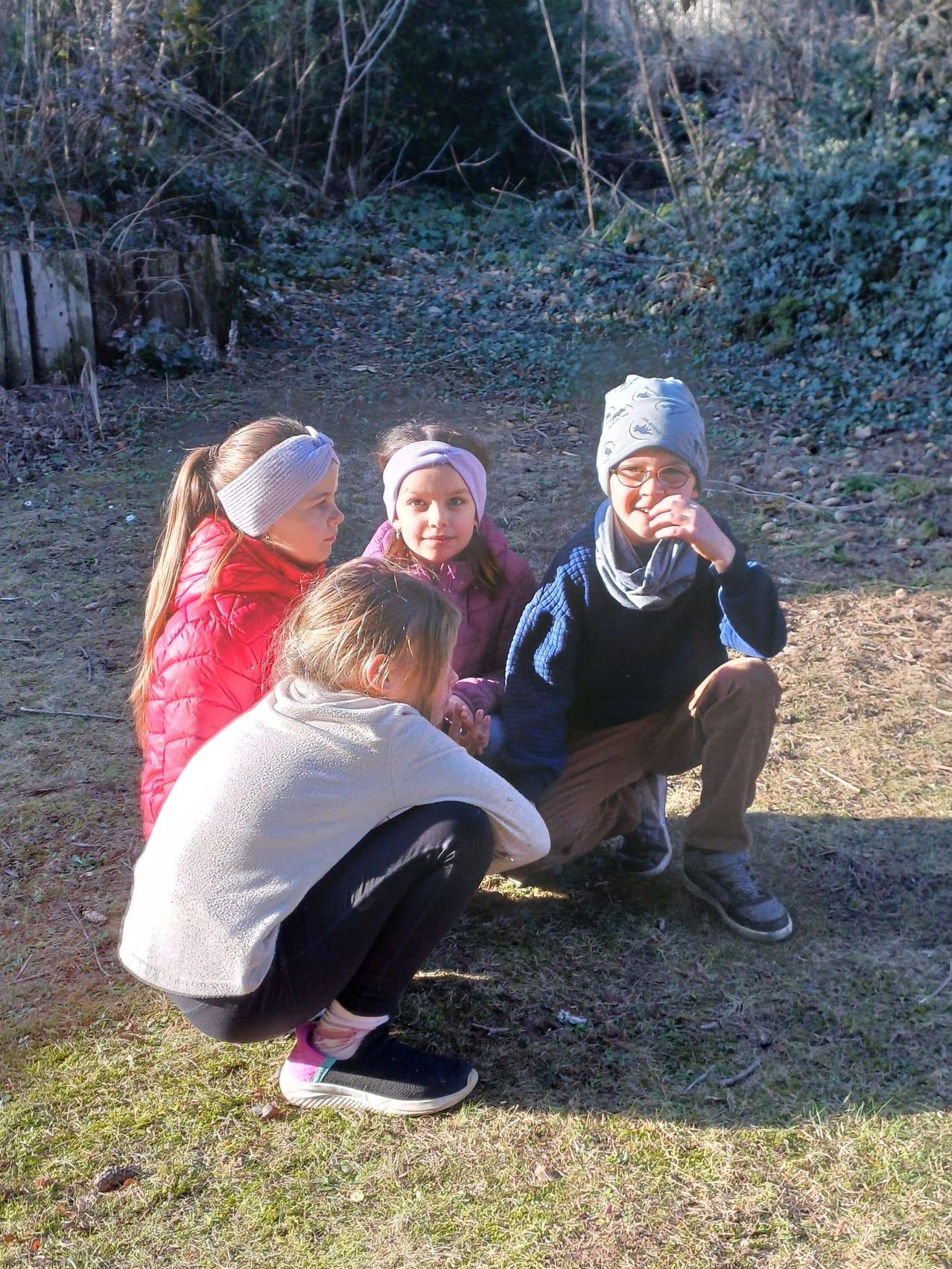 Four children are sitting on the ground, likely in a park. They are smiling and looking at something. The girl in gray is kneeling. Behind them is a wooden fence and some greenery.