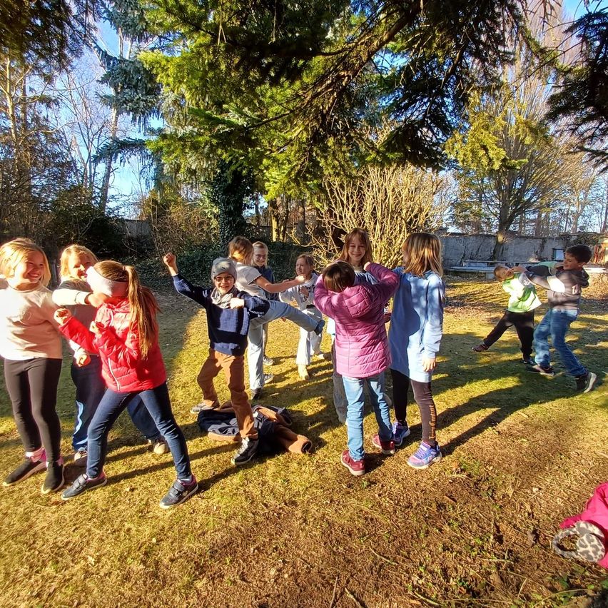 A group of children are playing outside on a sunny day. They are forming a circle and appear to be doing some group activity. Trees and a fence are in the background.