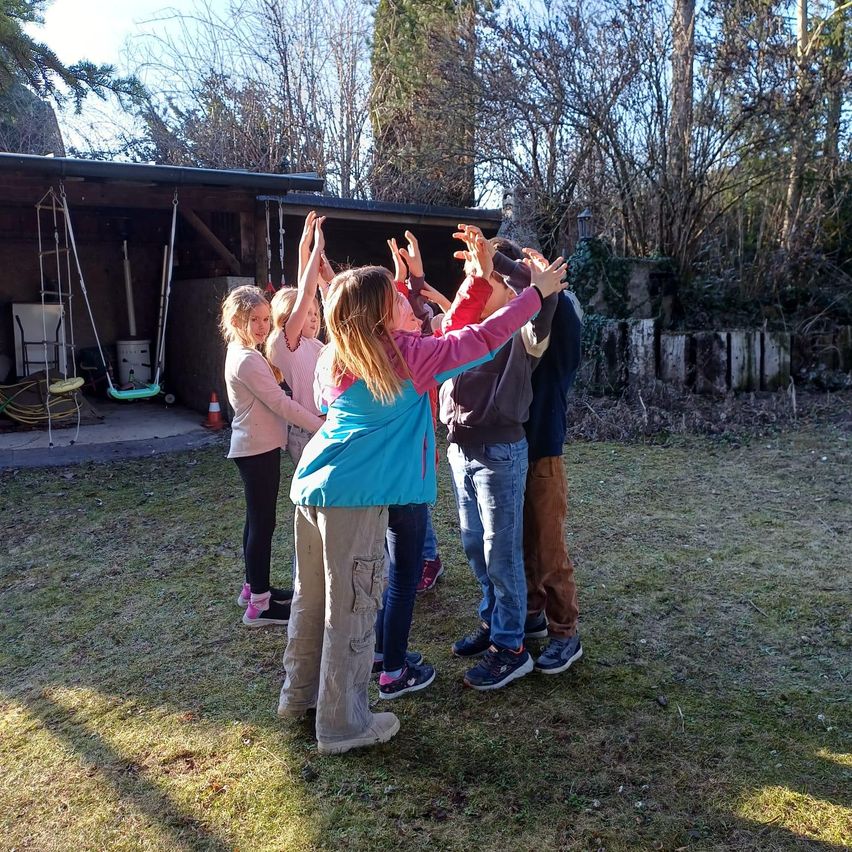 A group of children and adults are playing a game in a yard, surrounded by trees and a wooden fence.