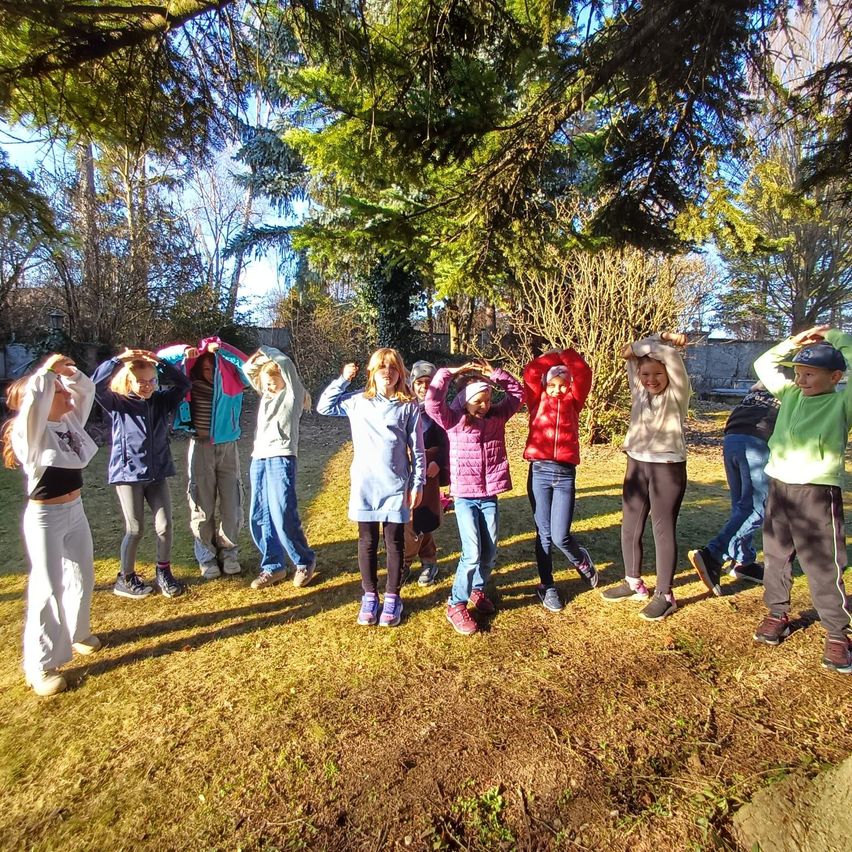 A group of children, dressed in various jackets and shoes, stand in a circle under a tree in a park. They are making hand gestures, possibly during a group activity.