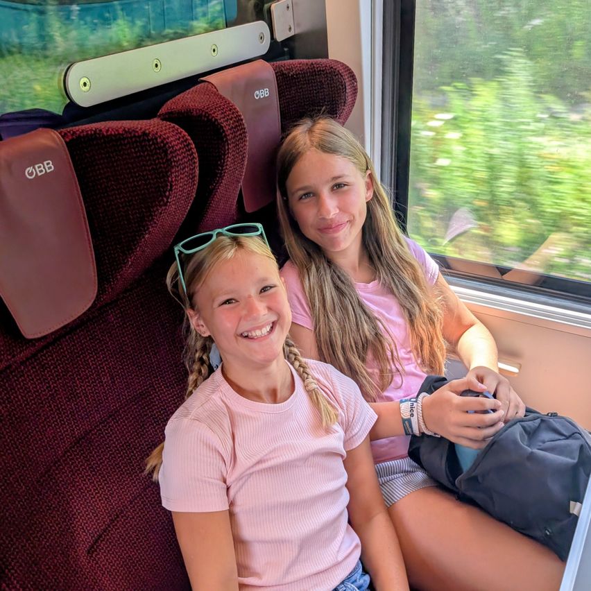 Zwei junge Mädchen sitzen in einem Zug, lächeln und schauen aus dem Fenster. Das Mädchen auf der linken Seite trägt eine Sonnenbrille auf dem Kopf und ein rosa Shirt. Das Mädchen auf der rechten Seite hält eine Tasche und trägt ein rosa Shirt.