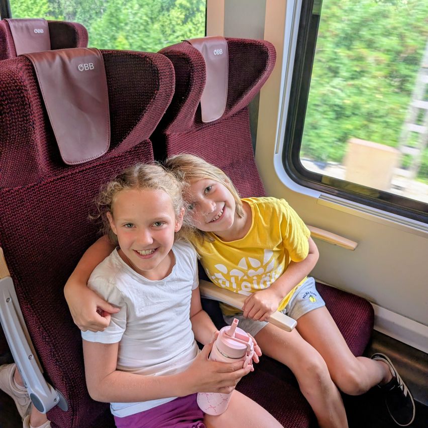 Zwei Mädchen lächeln in einem ÖBB-Zug mit grüner Landschaft im Hintergrund.