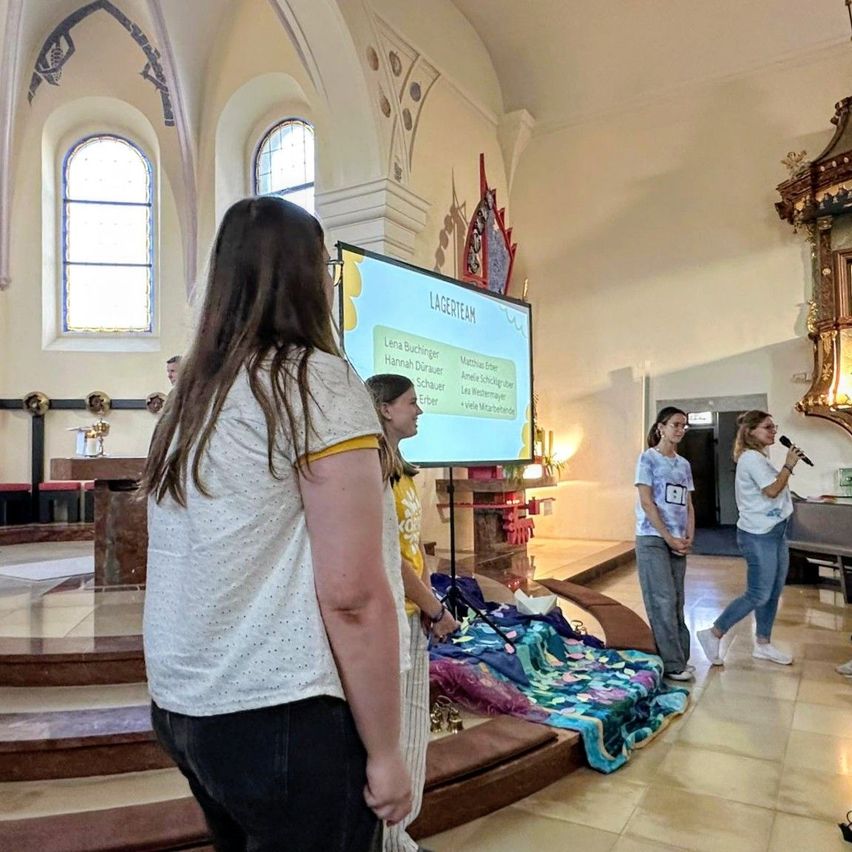 Eine Gruppe von Frauen versammelt sich in einer Kirche, wobei eine Frau in ein Mikrofon spricht. Eine Projektionsleinwand zeigt eine Liste von Namen an.