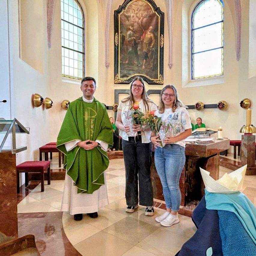 Drei Personen stehen in einer Kirche, eine in einem grünen Gewand, zwei halten Blumen. Hinter ihnen zieren ein Gemälde und Buntglasfenster die Wand.