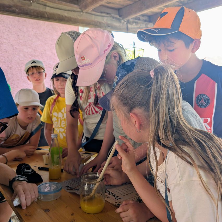 Eine Gruppe von Kindern versammelt sich um einen Tisch, wobei ein Mädchen mit einem Strohhalm aus einem Glas mit Orangensaft pustet. Sie tragen alle Mützen und scheinen an einer Aktivität beteiligt zu sein.