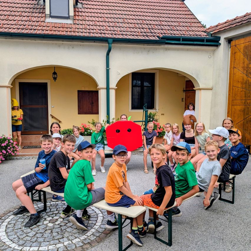 Kinder sitzen auf Bänken vor einem Haus mit einem roten Apfel-Kostüm