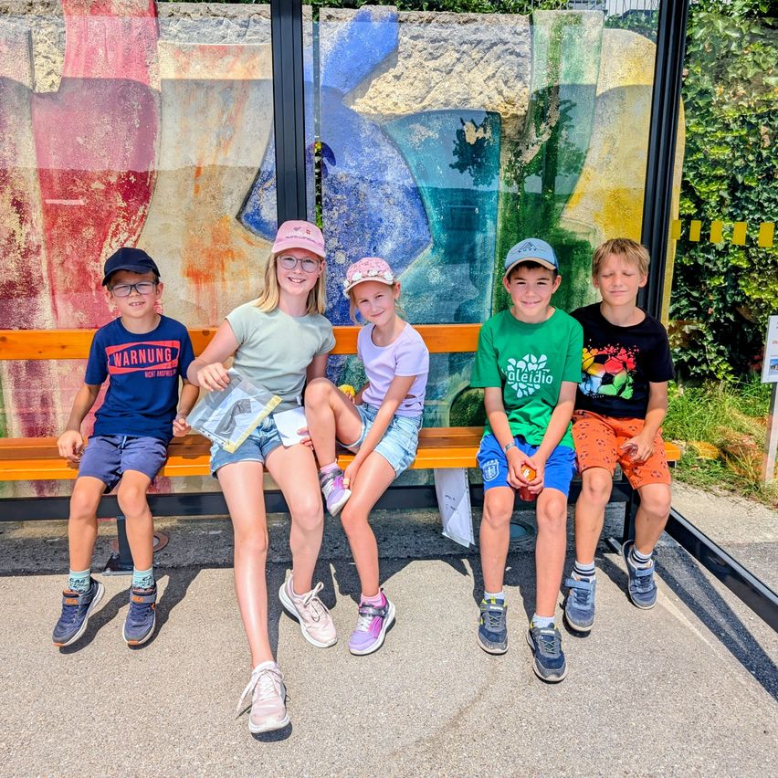Fünf Kinder sitzen auf einer Bank vor einer bunten Glaswand.