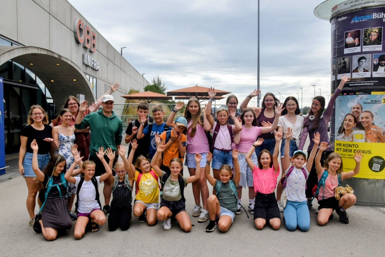 A group of children and adults pose for a photo outside a building with the word 'INFRA' on it. The children are wearing backpacks and some are wearing hats.