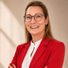Eine Frau in einem roten Blazer, weißem Hemd und Brille steht mit verschränkten Armen und lächelt für ein Foto.