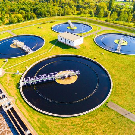 Ein Luftbild einer Kläranlage zeigt mehrere große, runde Tanks und ein zentrales Gebäude. Die Tanks sind mit dunklem Wasser gefüllt, und eine Brücke verbindet sie. Die Anlage ist von grünem Gras und Bäumen umgeben.