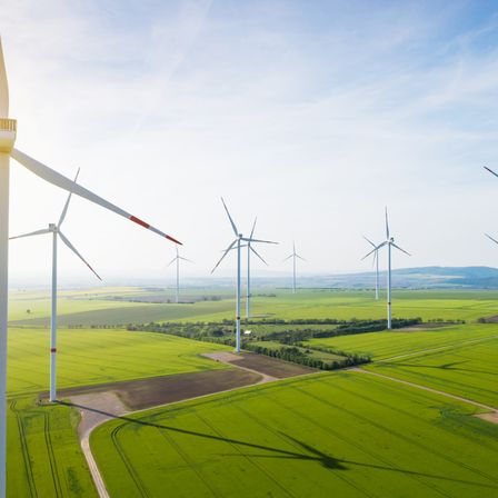 Ein Windpark mit mehreren Turbinen steht in einem weitläufigen grünen Feld unter einem klaren blauen Himmel mit einigen Wolken.