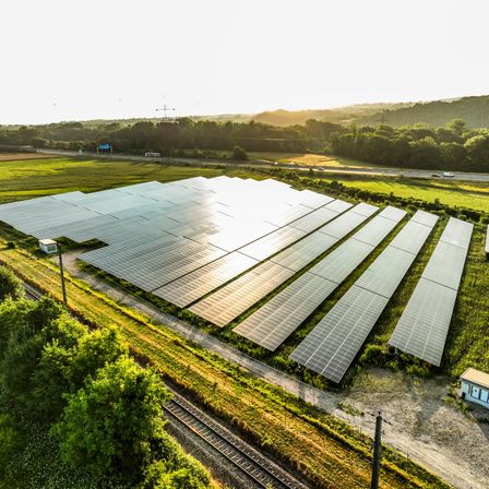 Luftaufnahme einer Solaranlage mit vielen Sonnenkollektoren, die das Sonnenlicht reflektieren, in einem grünen Feld mit einer Straße und Bergen im Hintergrund.