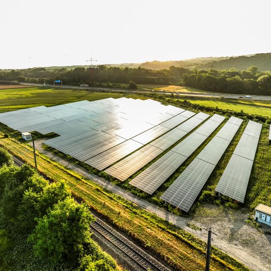 Luftaufnahme einer Solaranlage mit vielen Sonnenkollektoren, die das Sonnenlicht reflektieren, in einem grünen Feld mit einer Straße und Bergen im Hintergrund.