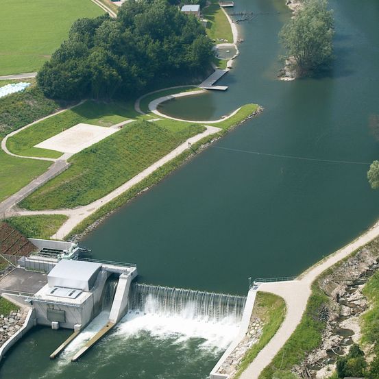 Luftaufnahme einer Staumauer mit durchfließendem Wasser, umgeben von grünem Gras und Bäumen, mit einem Haus in der Ferne.