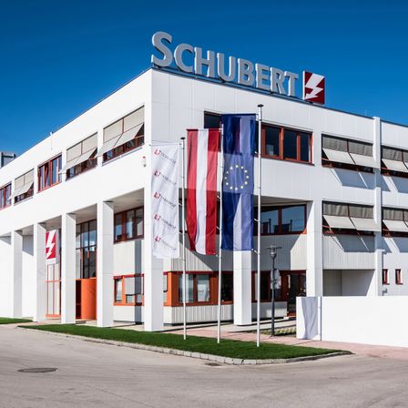 Ein modernes weißes Gebäude mit dem Namen Schubert oben drauf. Es hat Flaggen von Österreich, Frankreich und der Europäischen Union. Das Gebäude befindet sich in der Nähe einer Straße mit einem Grasbereich davor.