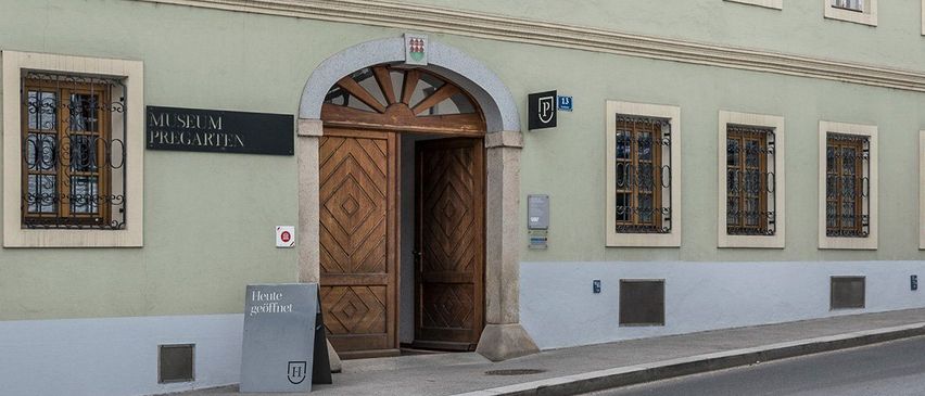 Ein hellgrünes Gebäude mit einem Ziegeldach hat mehrere Fenster und einen hölzernen Bogeneingang. Das Schild über dem Eingang steht 'Museum Pregarten'. Neben dem Eingang befindet sich ein schwarzes Schild mit einem Logo an der Wand.