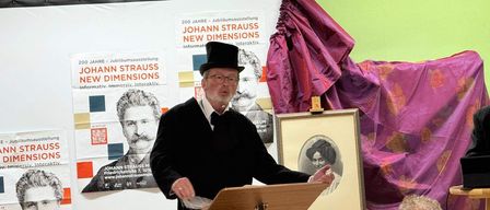 Ein Mann in formeller Kleidung steht an einem Podium und spricht zu einer Gruppe von Menschen. Hinter ihm befindet sich ein Banner zur 200-Jahr-Feier von Johann Strauss mit Porträts.