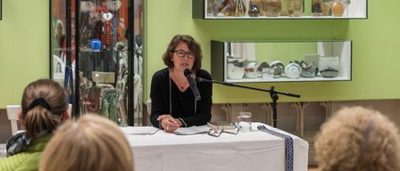 Eine Frau spricht in ein Mikrofon an einem Tisch, mit einem Glas Wasser und einem Buch vor ihr. Mehrere Menschen lauschen. Hinter ihr hält eine grüne Wand Glasdisplays mit verschiedenen Gegenständen.