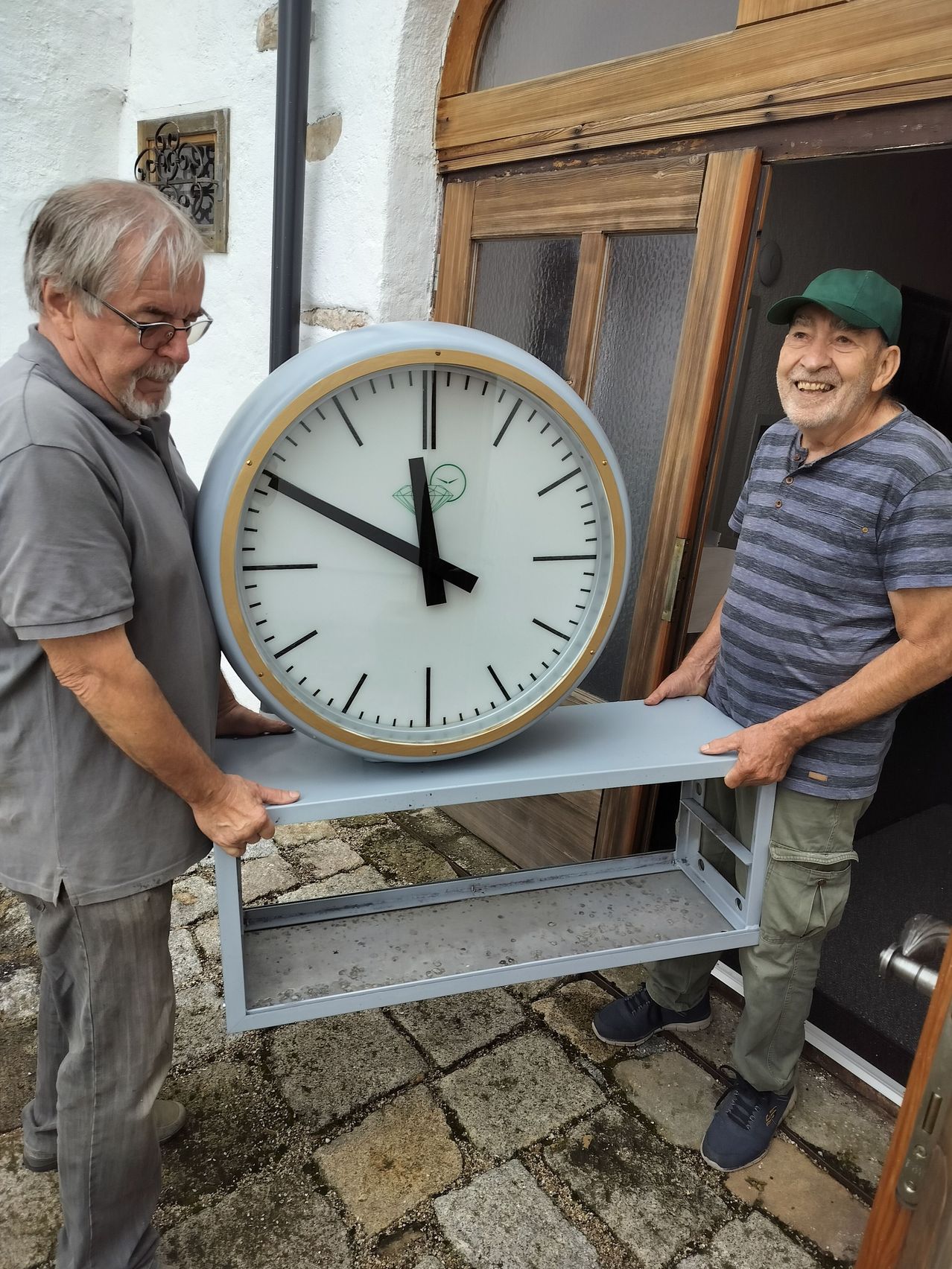 Zwei Männer halten eine große Uhr auf einer Plattform vor einem Gebäude. Die Uhr hat ein weißes Zifferblatt mit schwarzen Zeigern. Der Mann auf der linken Seite trägt eine Brille und ein graues Hemd, und der Mann auf der rechten Seite trägt eine grüne Mütze und ein gestreiftes Hemd.
