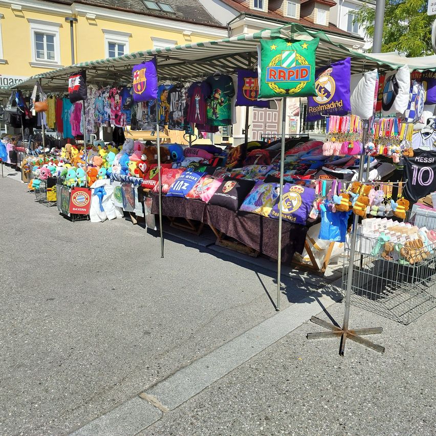 Ein Straßenmarkt mit verschiedenen Waren unter einem Zelt, darunter FC Barcelona und Real Madrid Trikots, Spielzeug und andere Artikel.