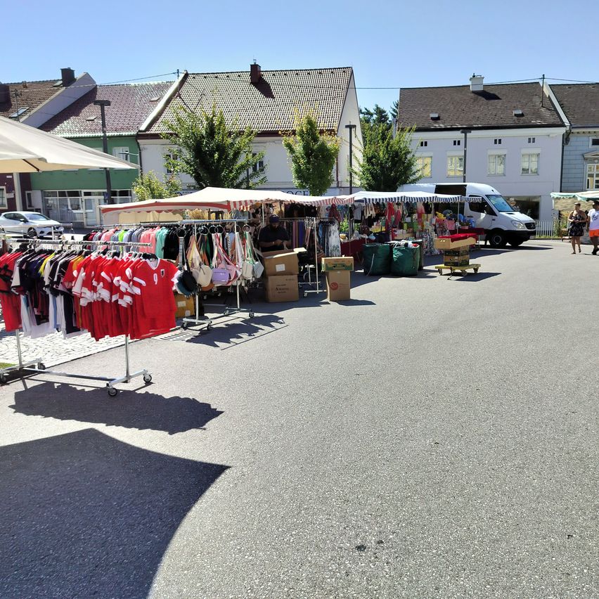 Ein Freiluftmarkt auf einem Stadtplatz mit Ständen, die verschiedene Artikel verkaufen, darunter Kleidung und Accessoires. Eine Person stöbert durch die Stände. Im Hintergrund befinden sich Gebäude und Bäume.