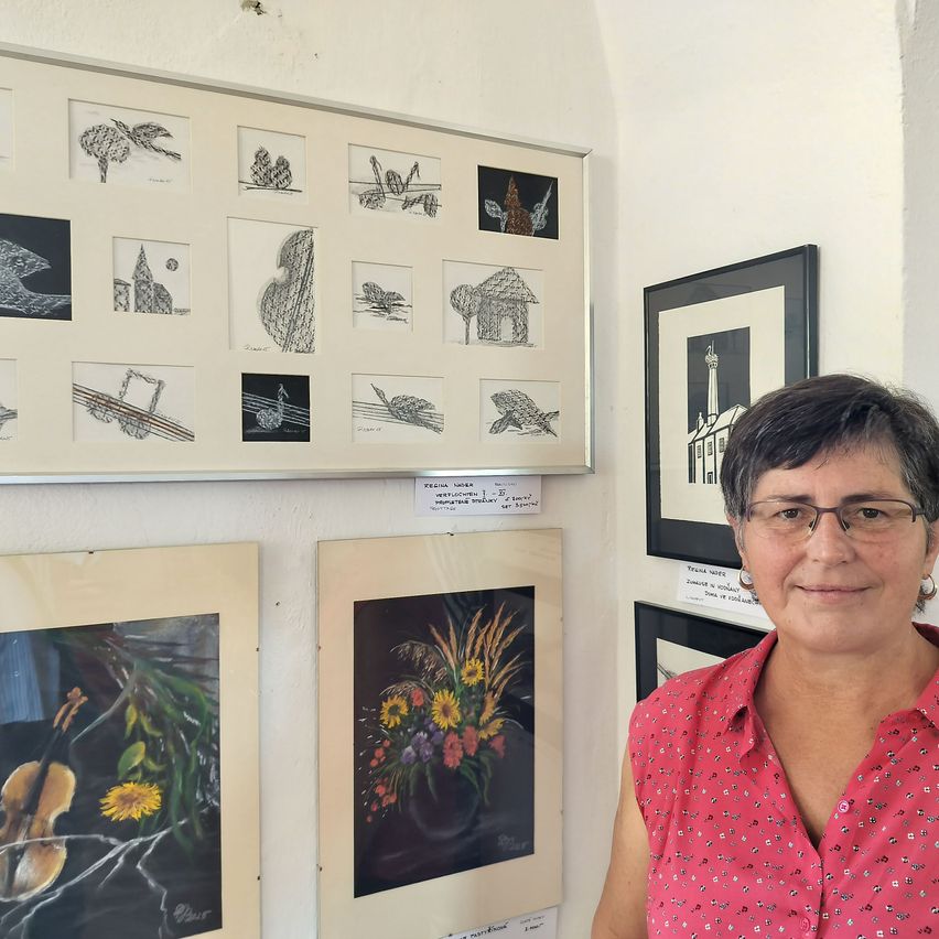 Eine Frau steht vor einer Wand, die mit verschiedenen gerahmten Kunstwerken geschmückt ist. Die Frau trägt eine Brille und ein rosafarbenes Oberteil. Die Gemälde variieren im Stil und zeigen Blumenarrangements, Musikinstrumente und abstrakte Designs.