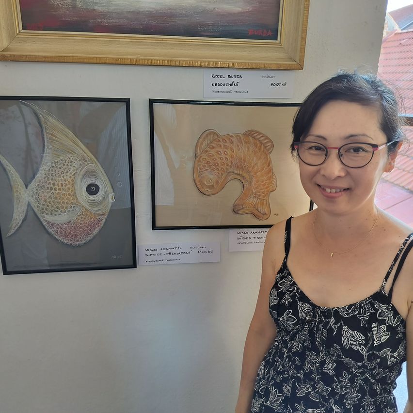 Eine Frau steht lächelnd vor einer Wand mit zwei gerahmten Bildern. Ein Bild zeigt einen Fisch, und das andere einen goldenen Fisch. Darüber befinden sich zwei weitere gerahmte Bilder.