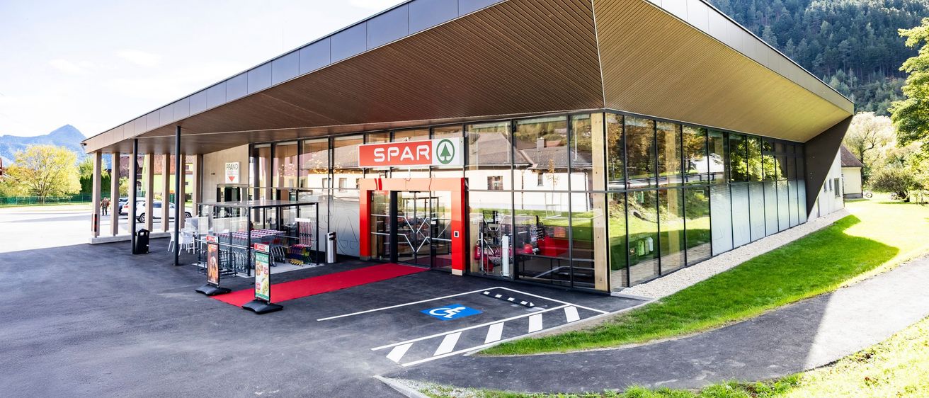Das Äußere eines modernen Spar-Supermarkts mit einem geneigten Dach und Glaswänden, umgeben von Gras und Bergen.