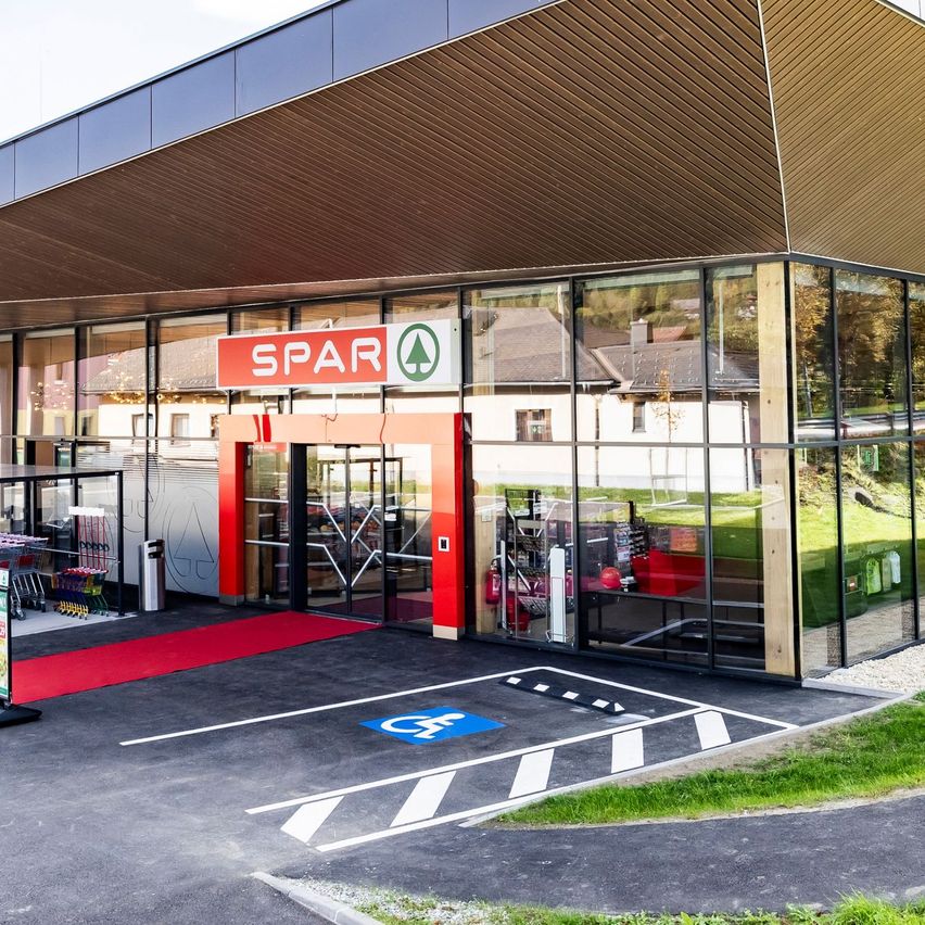 Das Äußere eines modernen Spar-Supermarkts mit einem geneigten Dach und Glaswänden, umgeben von Gras und Bergen.