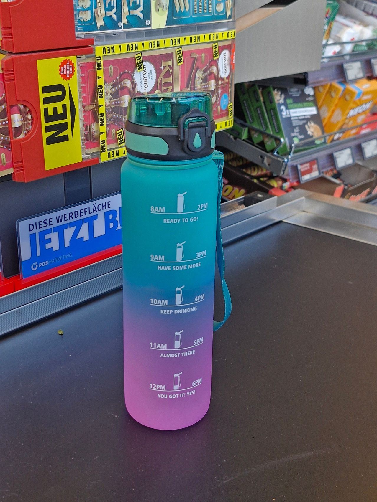 Eine pinke und blaue Wasserflasche mit einem Zeitplan darauf steht auf einer schwarzen Oberfläche. Dahinter ist ein verschwommener Hintergrund mit einem gelben Schild, das 'Neu' steht.