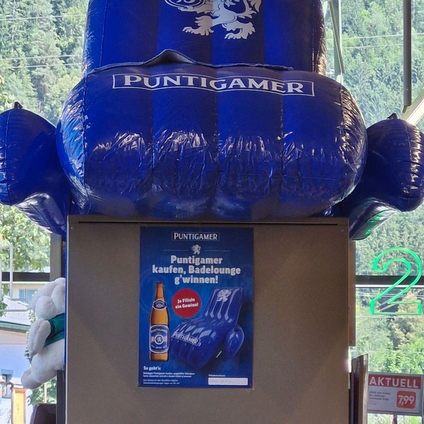Ein blauer aufblasbarer Stuhl mit dem Wort Puntigamer darauf, auf einem Karton mit einem Plakat, das Puntigamer-Bier bewirbt. Das Plakat zeigt eine Bierflasche und einen Liegestuhl.