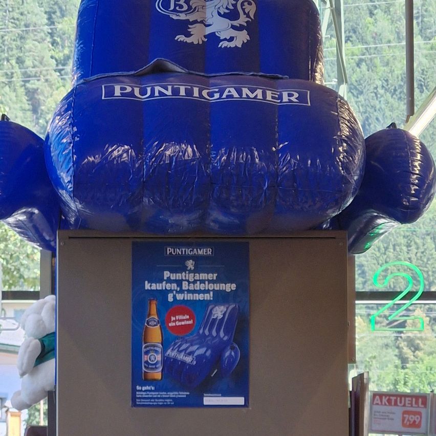 Ein großer aufblasbarer blauer Stuhl mit dem Wort Puntigamer darauf. Darunter wirbt ein Poster für Puntigamer-Bier mit einer Flasche und einer Pool-Liege. Ein weißes Kuscheltier ist links.