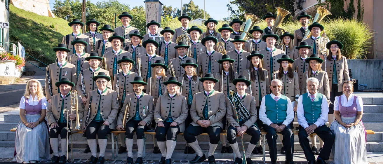 Ein Blasorchester in traditioneller Kleidung posiert für ein Gruppenfoto. Sie sitzen auf Steinstufen mit Musikinstrumenten vor einer Kulisse aus Grünflächen und einem historischen Denkmal.