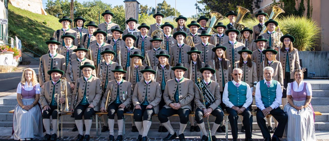 Ein Blasorchester in traditioneller Kleidung posiert für ein Gruppenfoto. Sie sitzen auf Steinstufen mit Musikinstrumenten vor einer Kulisse aus Grünflächen und einem historischen Denkmal.