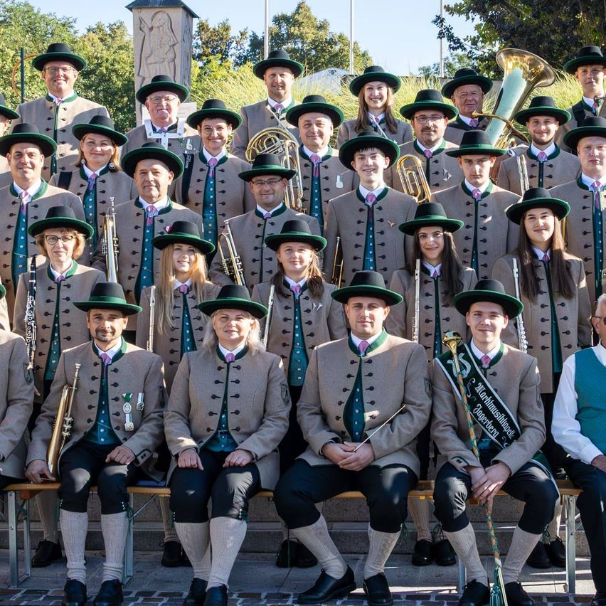 Ein Blasorchester in traditioneller Kleidung posiert für ein Gruppenfoto. Sie sitzen auf Steinstufen mit Musikinstrumenten vor einer Kulisse aus Grünflächen und einem historischen Denkmal.