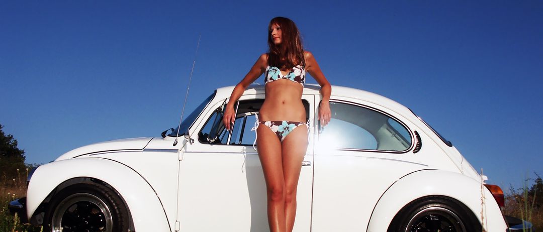 Eine Frau in einem Bikini lehnt an einem weißen Oldtimer in einem offenen Feld unter einem klaren blauen Himmel.