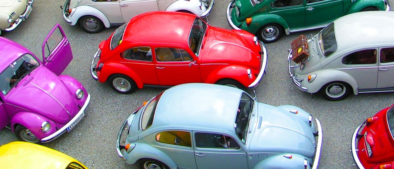 Ein Parkplatz voller klassischer Autos. Die Autos sind in verschiedenen Farben und in Reihen geparkt. Einige der Autos haben Gepäck auf dem Dach. Eine Person steht in der Nähe eines roten Autos.