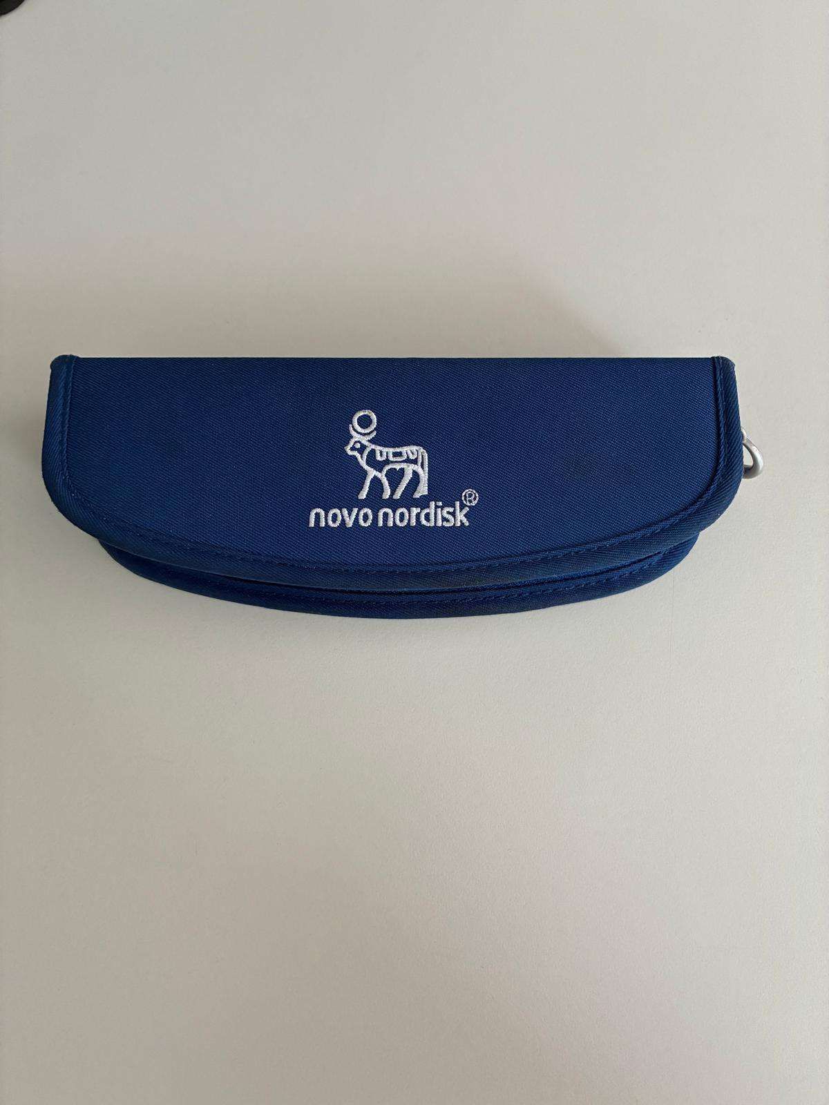 Eine blaue Tasche mit einem weißen Novo Nordisk Logo, das eine stilisierte Kuh und den Firmennamen zeigt. Die Tasche ist zum Transportieren und Schützen von Gegenständen gedacht.