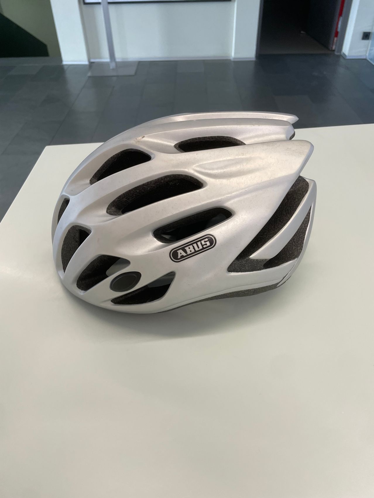 Ein silberner Fahrradhelm mit schwarzen Akzenten und dem Wort 'ABUS' in weiß auf der Vorderseite wird auf einer weißen Oberfläche ausgestellt.